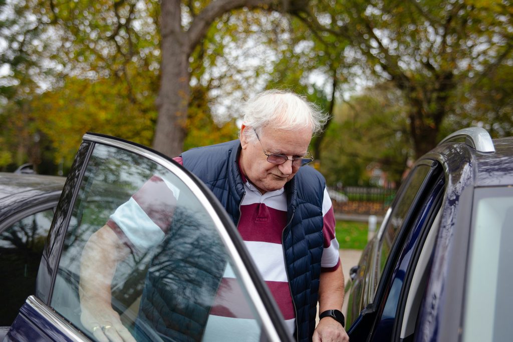 Smiling senior woman being helped into the front seat of a car by a caregiver on a quiet, tree-lined street